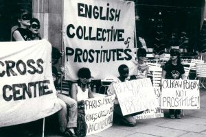 Black and white photo of a English Collective of Prostitutes banner with people stood and kneeling with signs