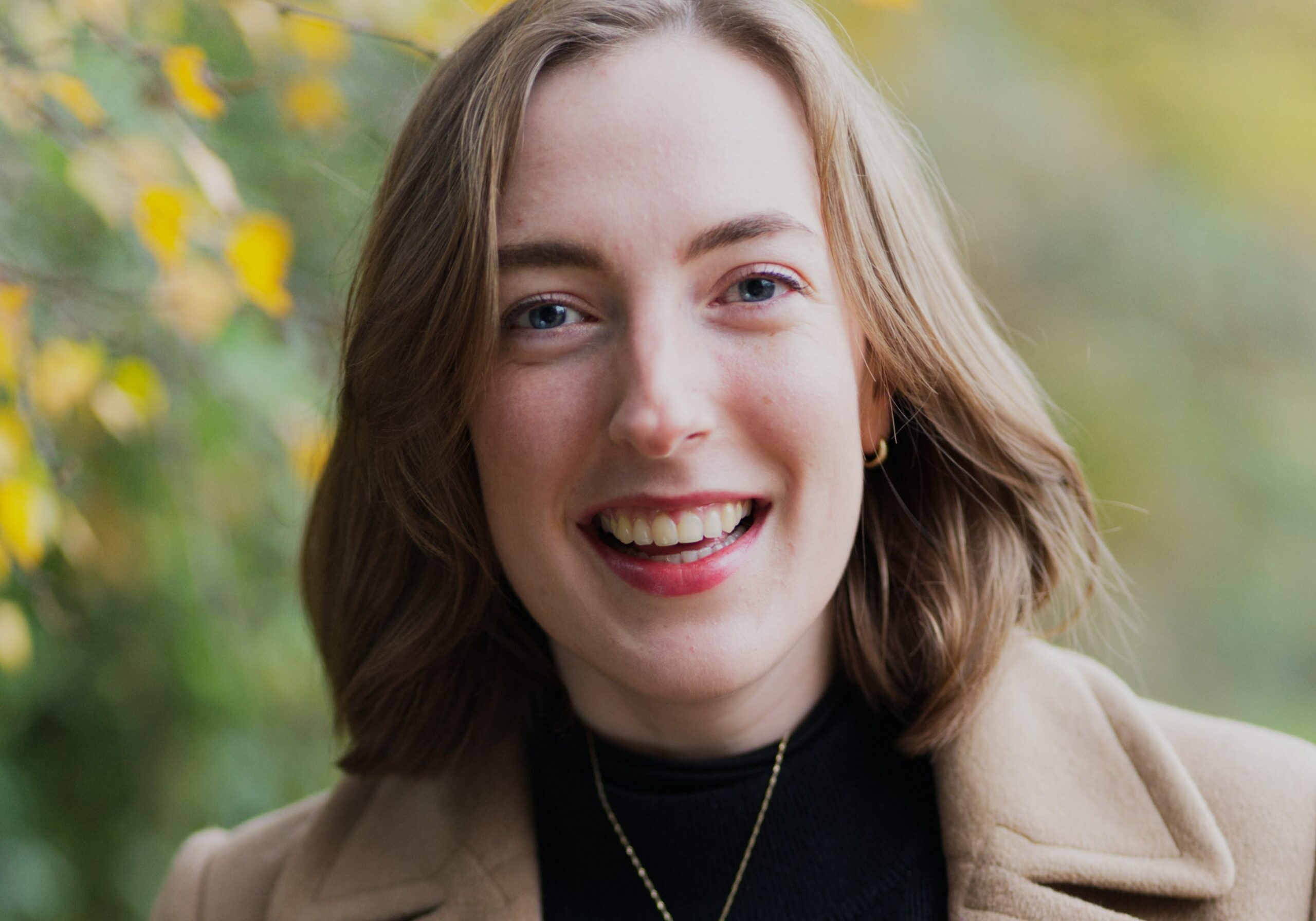 A photo of Lauren McLay smiling into camera dressed smartly with a leafy, autumnal background